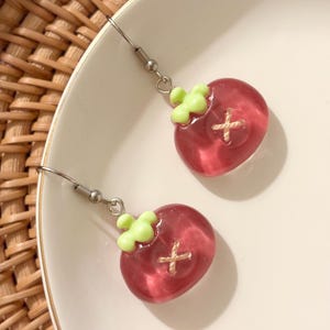 May include: A pair of dangle earrings featuring a translucent red fruit design with a light green stem and a stitched cross detail. The earrings are displayed on a white plate.