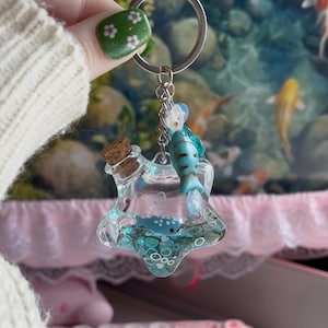 May include: A star-shaped clear resin keychain filled with liquid, small blue fish charms, and white circular accents. The keychain features a cork stopper at the top, a silver metal ring, and a dangling blue fish charm attached to the silver chain.