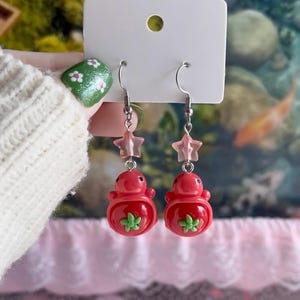 May include: A pair of dangle earrings featuring red strawberry-shaped charms with small green leaf details. Each earring hangs from a silver-toned metal hook and includes a translucent pink star-shaped bead positioned above the strawberry charm.