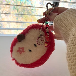 May include: A plush, red and white hedgehog keychain with a heart-shaped clasp. The keychain features a pink star and a brown ear. Decorative charms include a small apple, stars, and hearts.