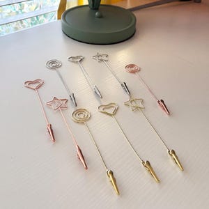 May include: A collection of decorative metal clips in various shapes, including hearts, stars, and spirals. The clips are in silver, gold, and rose gold colors. Each clip has a small alligator clip at the bottom.