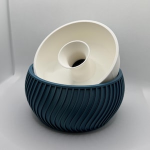 May include: A two-piece decorative bowl set. The base is a dark teal with a ribbed, curved design. The top piece is a white, circular bowl with a central, cylindrical element. The set is designed for decorative purposes.