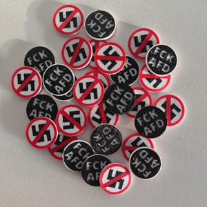 Könnte beinhalten: Eine Ansammlung kleiner, runder Aufnäher in Schwarz und Weiß. Einige Aufnäher zeigen ein rotes Symbol mit einem schwarzen Symbol, das durchgestrichen ist. Andere haben den Text "FCK AFD" in Weiß.