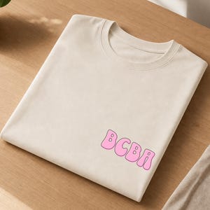 Peut inclure: Un t-shirt beige à manches courtes avec une encolure ronde. Le vêtement présente les lettres BCBA imprimées dans une police rose, arrondie et de style bulle sur le côté inférieur droit. Le t-shirt est plié, montrant une texture de tissu lisse.