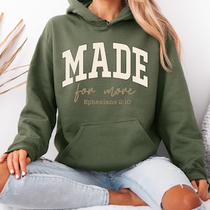 Könnte beinhalten: Olivgrüner Kapuzenpullover mit dem Text "MADE for more" in fetter, cremefarbener Schrift. Darunter steht "Ephesians 2:10" in kleinerer, kursiver Schrift. Der Hoodie hat eine Vordertasche und eine Kapuze mit Kordelzug.