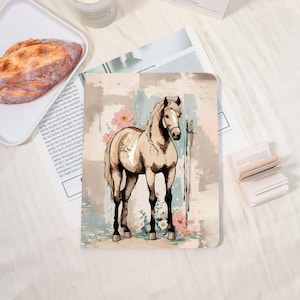 May include: A beige horse illustration on a notebook cover. The horse is standing with a bridle and a sword. Pink flowers and a watercolor background in beige, blue, and pink complete the design. The notebook is on a white surface.