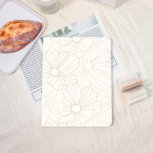 May include: A cream-colored notebook with a floral design. The cover features a pattern of large, outlined flowers in a soft, muted green. The notebook is rectangular and appears to be lying on a white surface, with other objects in the background.