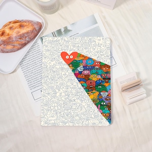 May include: A notebook with a whimsical design. The cover features a white background with a pattern of small, cartoonish faces. A red heart with a smiling face is at the top, with a colorful, triangular section filled with more cartoon characters.