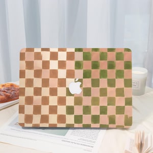 May include: A laptop case with a checkered pattern in shades of brown, green, and pink. The Apple logo is visible in the center. The case is on a table with a pastry, a candle, and a newspaper.