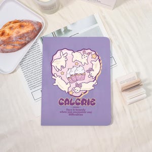 May include: A purple notebook with a heart-shaped design featuring white rabbits, cupcakes, and stars. The word "CALCRIC" is printed below the design, along with the text "Face it bravely when you encounter any difficulties."