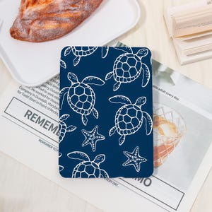 May include: A navy blue coaster featuring a white outline pattern of sea turtles and starfish. The coaster is square and sits on a white surface. The design is repeated across the coaster's surface.