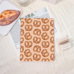 May include: A notebook with a repeating pattern of brown pretzels on a peach background. The notebook is open, and the pages are visible. The image also shows a loaf of bread, a candle, and a stack of small white notepads.
