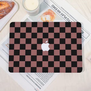May include: A laptop skin featuring a checkerboard pattern in black and a muted reddish-brown. The Apple logo is centered on the skin. The skin is on a white surface with a newspaper and other items in the background.