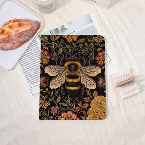 May include: A notebook with a black background featuring a large bumblebee illustration. The bee is centered, with yellow and black stripes. The background is filled with floral patterns in shades of red, yellow, and green.