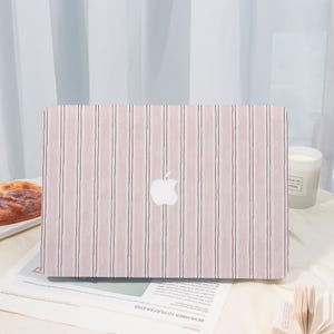 May include: A laptop with a pink and white striped design. The Apple logo is centered on the cover. The laptop is on a table with a pastry, a candle, and a newspaper.