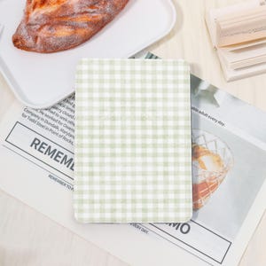 Könnte beinhalten: Ein rechteckiger Untersetzer mit grün-weißem Vichy-Muster. Der Untersetzer liegt auf einer Zeitung, auf der das Wort "REMEMBER" zu sehen ist. Ein Brotlaib liegt auf einem weißen Teller.