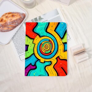 May include: A colorful notebook with a vibrant abstract design. The cover features a swirling spiral in shades of yellow, orange, red, and blue, outlined in black. The notebook is on a white surface.