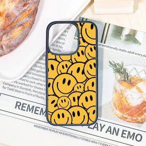 May include: A yellow phone case with a black border, featuring a repeating pattern of smiling faces. The case is on a white surface with a pastry and a glass of liquid with a sprig of rosemary.