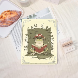 Könnte beinhalten: Eine skurrile Illustration einer Froschfigur mit einem Pilzhut, der in einem roten Buch liest. Der Frosch ist von grünem Laub umgeben, vor einem cremefarbenen Hintergrund mit dekorativen Rändern. Das Bild hat eine Vintage-Ästhetik.