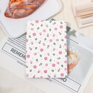 May include: A white notebook with a floral pattern of pink flowers and green leaves. The notebook is open on a newspaper with the word "REMEMBER" visible. A pastry is on a white plate in the background.