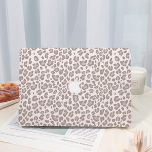 May include: A laptop hard shell case featuring a repeating leopard print pattern in shades of light gray and beige. The design covers the entire surface of the case, which includes a central cutout for the Apple logo on the laptop lid.