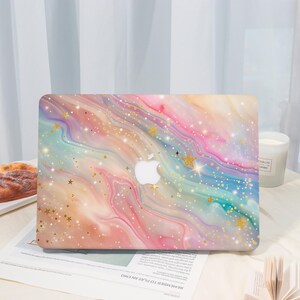 May include: A laptop case with a colorful, swirling design in shades of pink, blue, yellow, and purple. The case features a white Apple logo and is decorated with gold stars and glitter. The case is on a white surface.