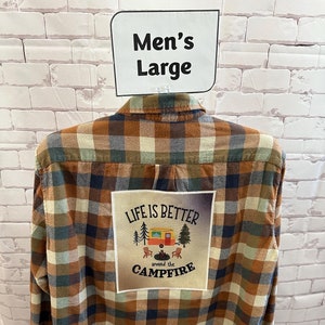 May include: A brown, blue, and beige plaid flannel shirt with a patch on the back that says "Life is better around the campfire" with a small camper and trees in the background.