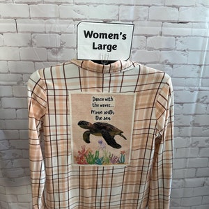 May include: A long-sleeved plaid shirt in peach, cream, and brown tones. The back features a rectangular patch with a sea turtle illustration and the text "Dance with the waves... Move with the sea." The shirt is labeled "Women's Large."