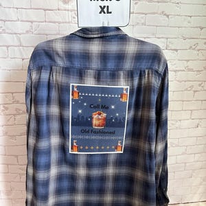 May include: Blue and gray plaid flannel shirt with a rectangular patch featuring an Old Fashioned cocktail and the text "Call Me Old Fashioned." The shirt has a "Men's XL" tag.