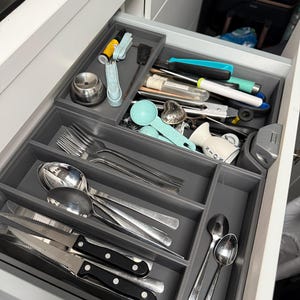 Könnte beinhalten: Geöffnete Schublade mit grauen Trennwänden, gefüllt mit Besteck und Küchenutensilien. Enthält Gabeln, Löffel, Messer und verschiedene Werkzeuge. Die Schublade ist Teil eines weißen Schranks.
