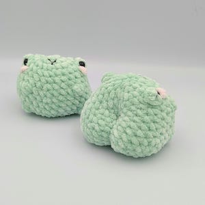 May include: Two green crocheted frog plushies with pink accents on their faces.