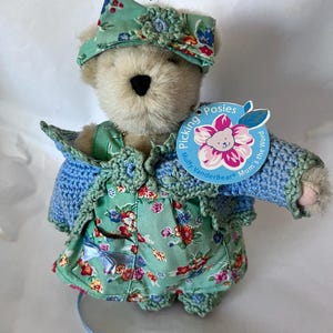 May include: A plush teddy bear wearing a floral print dress, a blue crocheted sweater, and a matching hat. The bear has a tag that reads "Picking Posies, Mum's the Word" with a flower graphic. The outfit features shades of blue, green, and red.