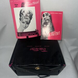 May include: A black vintage travel case with a gold clasp and handle, accompanied by two pink and white boxes. The boxes feature images of hairstyles and the text "Tovar Tresses". One box displays the price "25¢".