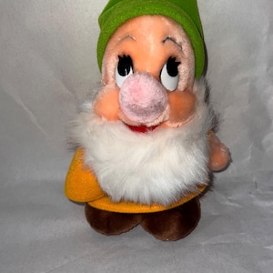 May include: A plush doll of the character Dopey from Snow White. The doll has a peach-colored face, a large pink nose, and a white furry beard. He wears a green hat, a yellow shirt, and brown shoes. The doll is a collectible item.