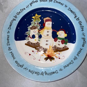 Pode incluir: Um prato redondo de cerâmica com uma cena de inverno. O prato apresenta três bonecos de neve em volta de uma fogueira, com uma árvore de Natal e o texto "Gather 'round for S'mores". A borda é azul clara com texto.