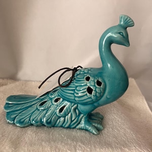 May include: A turquoise ceramic peacock figurine with a detailed tail and openwork design. The peacock has a long, curved neck and a decorative crest. A brown string is attached to the figurine.