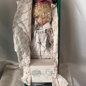 May include: A Knowles Dolls doll in its original packaging. The doll has blonde curly hair and is wearing a white dress with floral accents. The box is green and contains tissue paper with the Knowles Dolls logo.