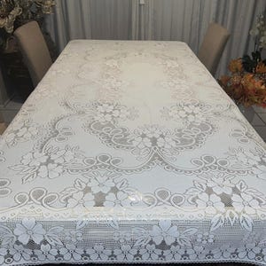 May include: A white rectangular tablecloth with a floral lace design. The tablecloth is spread over a wooden table, with two chairs visible on either side. The lace pattern features flowers and intricate details.