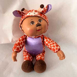 May include: A Cabbage Patch Kid doll wearing a giraffe costume. The costume is orange with brown spots and a purple belly. The doll has brown eyes, rosy cheeks, and brown hair. The doll's feet are brown.