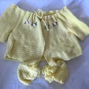 Könnte beinhalten: Hellgelbes Strick-Babyset, bestehend aus einer Strickjacke und zwei passenden Bloomers. Die Strickjacke hat einen Kordelverschluss und ist mit kleinen gestickten Blumen verziert. Das Set ist aus weichem, strukturiertem Garn gefertigt.