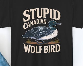 May include: Black t-shirt featuring a graphic of a loon with the text "STUPID CANADIAN WOLF BIRD" in a vintage style. The bird is depicted in shades of blue, gray, and white. The shirt is displayed on a white surface with a pair of blue jeans.