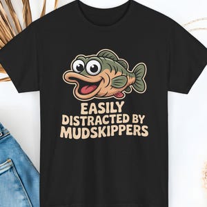May include: Black t-shirt featuring a cartoon fish with the text "EASILY DISTRACTED BY MUDSKIPPERS" in a playful font. The fish is green, tan, and pink with a smiling expression. A casual tee for fishing enthusiasts.