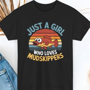 May include: Black t-shirt with a graphic featuring a cartoon fish and the text "JUST A GIRL WHO LOVES MUDSKIPPERS." The design includes a sunset background with blue, orange, and yellow hues. The fish is red and brown.