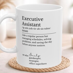 May include: White ceramic mug with the text "Executive Assistant" and a definition. The definition describes the role as someone who manages schedules, solves problems, and saves the day. The mug also includes the name "Emily" and other job titles.