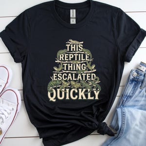 May include: A black short-sleeved t-shirt featuring a graphic print of various green lizards arranged in a tree-like shape. The text "THIS REPTILE THING ESCALATED QUICKLY" is printed in a light cream color, integrated among the lizard illustrations.
