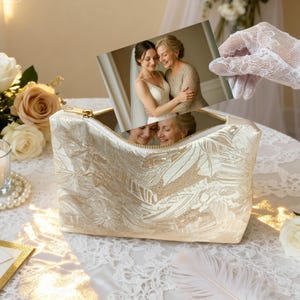 May include: A cream-colored cosmetic bag with a gold zipper and a floral pattern. The bag is open, revealing a photograph of two women embracing. The photo is held by a gloved hand. The scene is set on a lace-covered table.