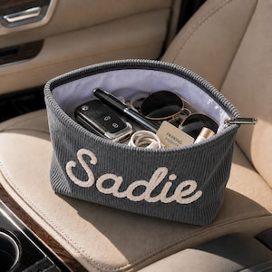 May include: Gray corduroy car organizer with the name "Sadie" embroidered in white. The organizer contains sunglasses, a pen, car keys, a charging cable, and a parking priority card. The organizer is sitting on a car seat.
