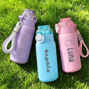 Puede incluir: Tres botellas de agua aisladas en lavanda, azul claro y rosa, cada una con una tapa y una correa de transporte a juego. La botella lavanda dice "Olivia", la botella azul claro dice "Sophia", y la botella rosa dice "Luna" con un dibujo de pingüino.