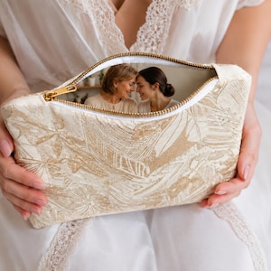 May include: A beige cosmetic bag with a gold zipper and a floral pattern. The bag is open, revealing a photograph of two women. The bag is held by a person wearing a white robe.