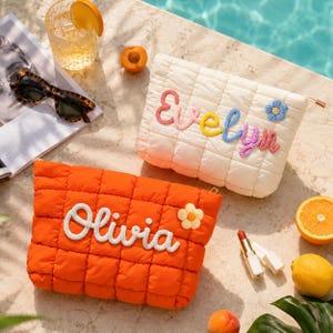 May include: Two rectangular quilted pouches, one cream and one orange, featuring embroidered names. The cream pouch displays the name Evelyn in multicoloured yarn with a blue flower, while the orange pouch features the name Olivia in white yarn with a yellow flower. Both have a zipper closure.